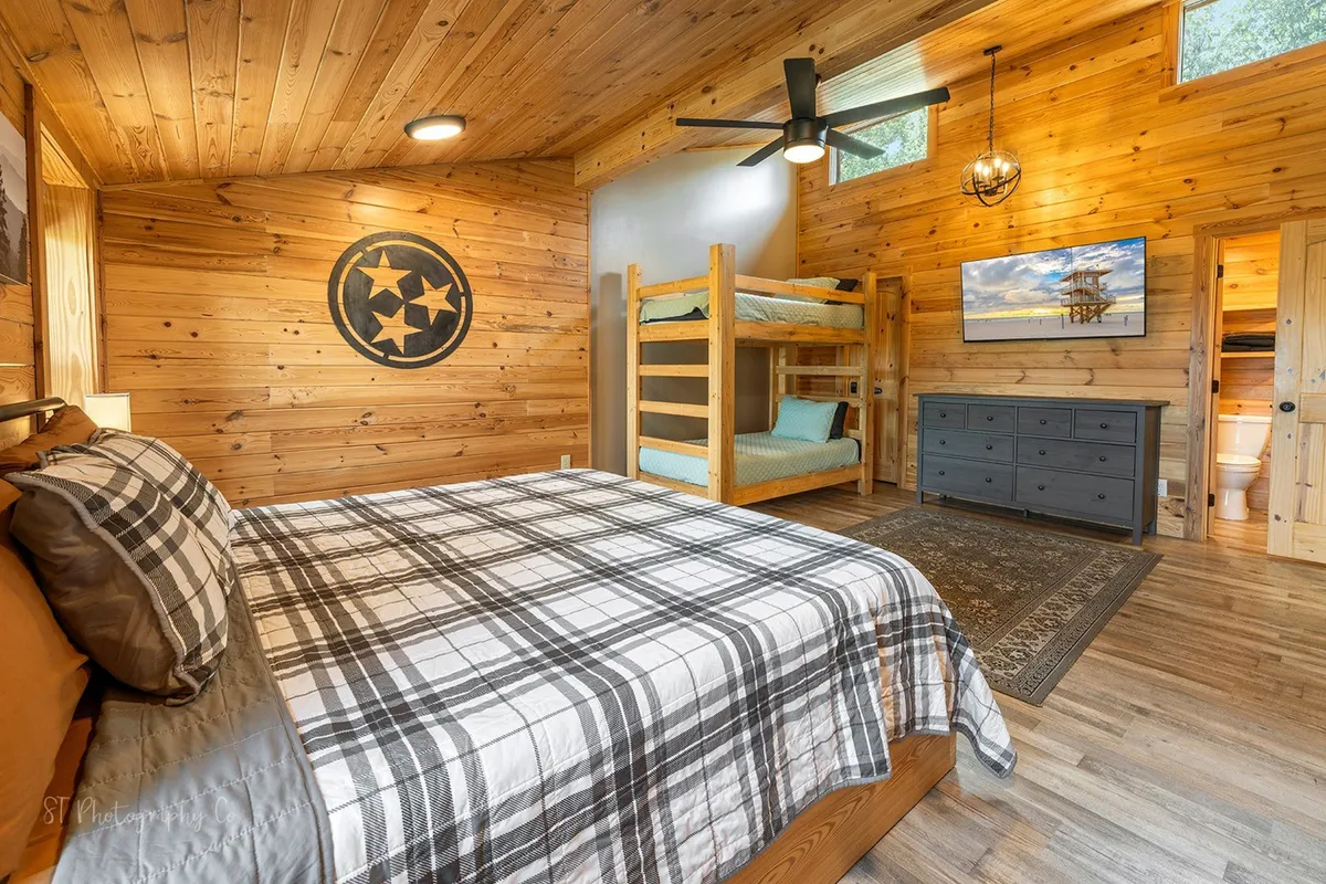 This image features a cozy bedroom with a king bed adorned in plaid bedding, a set of bunk beds ideal for kids or friends, wood-paneled walls, and a wall-mounted TV. The warm lighting and inviting atmosphere make it ideal for restful nights after days of adventure.