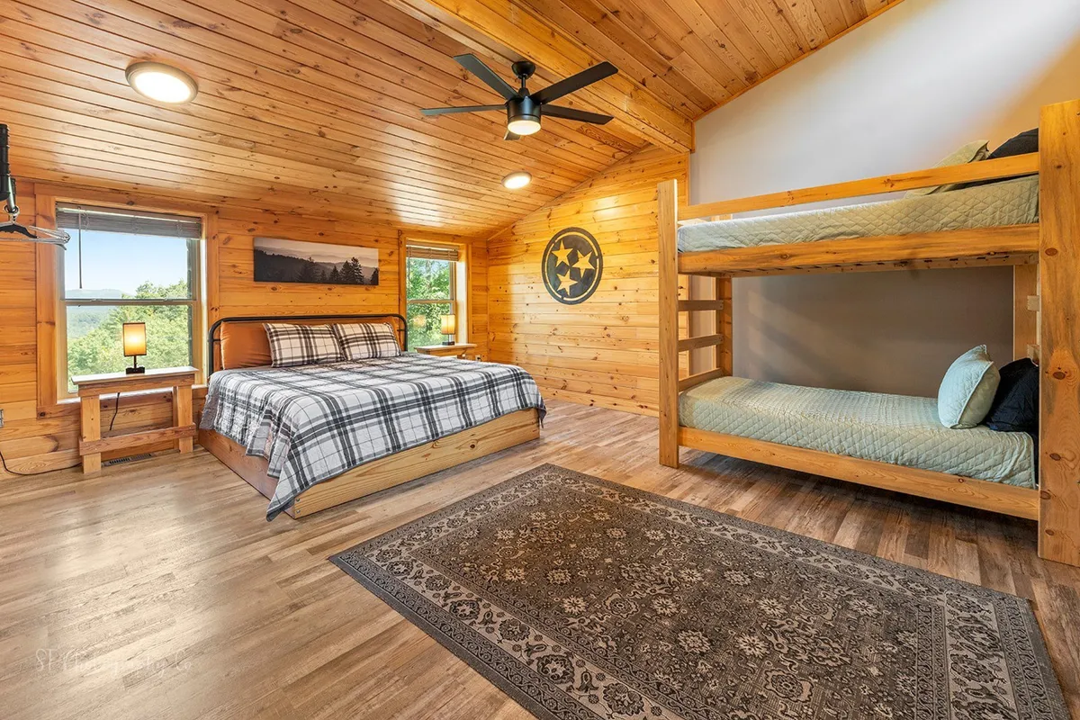 A welcoming bedroom features a queen bed and a bunk bed, with rustic wooden accents, a soft rug, and a window that lets in natural light. The setting is comfortable, relaxed, and ideal for family stays.