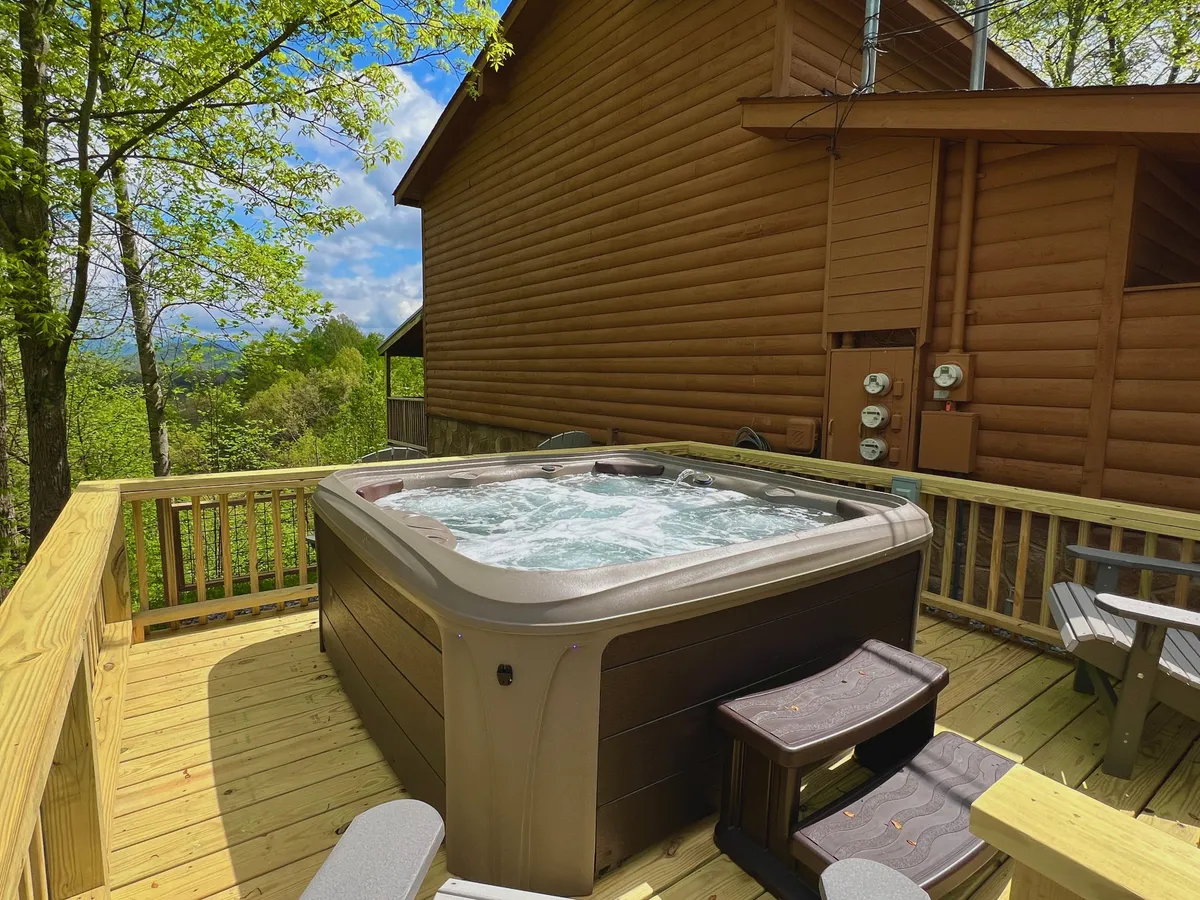 This image shows a spacious deck surrounded by lush trees, with a hot tub as the central feature. The hot tub sits beneath open sky, inviting guests to relax while taking in the natural mountain scenery. There is ample space for friends to gather and enjoy the outdoors together.
