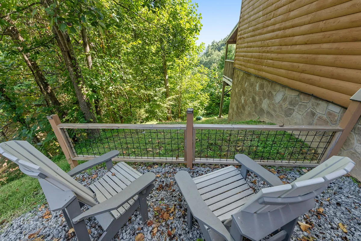 A row of adirondack chairs sits on a wooden porch, surrounded by natural landscaping and the rustic wooden siding of the cabin. The scene feels peaceful and welcoming, emphasizing connection to nature and space for group gatherings.