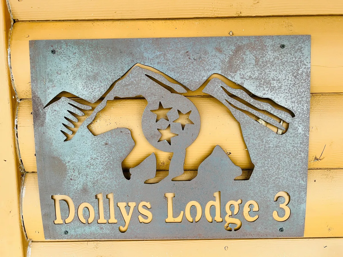 This image features a metal sign with a bear silhouette and mountain outline, along with the text 'Dollys Lodge 3.' The rustic sign is set against a natural backdrop, evoking a sense of adventure and welcoming families to the lodge.