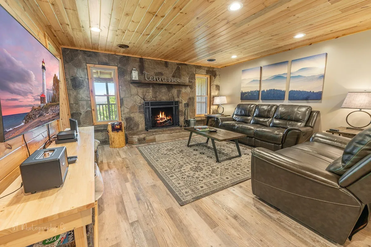 A spacious living room featuring a prominent wood-burning fireplace as the centerpiece, surrounded by large leather seating. Warm wooden accents and inviting artwork adorn the space, with a plush rug pulling the room together. Large windows offer glimpses of the natural surroundings, filling the room with light and creating a cozy, welcoming atmosphere.