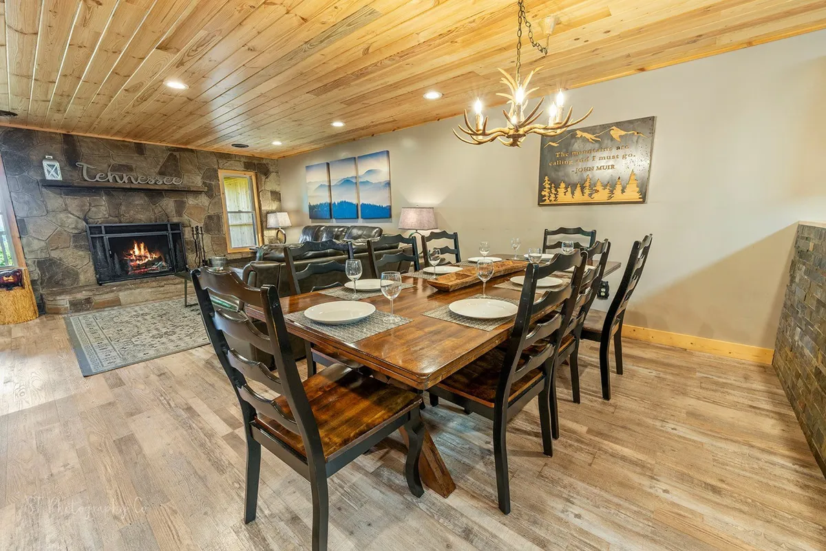 This image highlights a cozy dining area featuring a rustic dining table, a stone fireplace, a chandelier, and scenic views through the windows. The setting is inviting and open, with hints of the outdoors visible beyond the glass, emphasizing a seamless transition between indoor comfort and the natural surroundings.