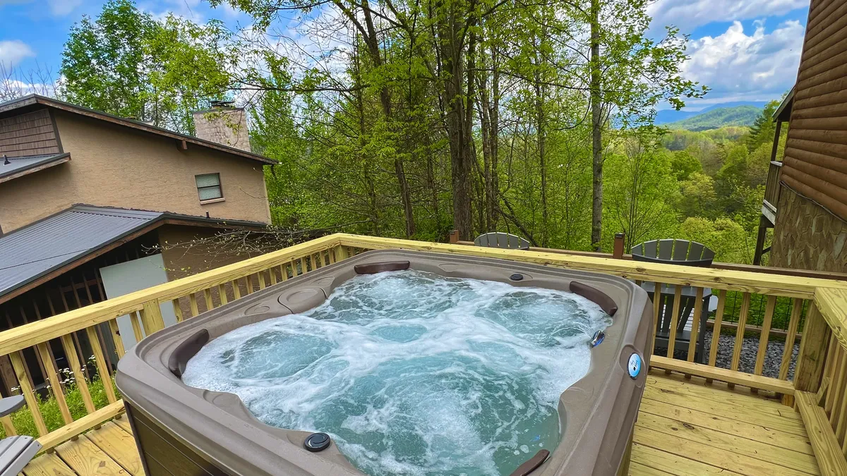 The image showcases the exterior of a cabin-style townhouse condo surrounded by lush greenery, with a private hot tub on the deck overlooking sweeping mountain views. The tranquil setting invites relaxation, with seating areas perfect for enjoying morning coffee or unwinding under the stars.