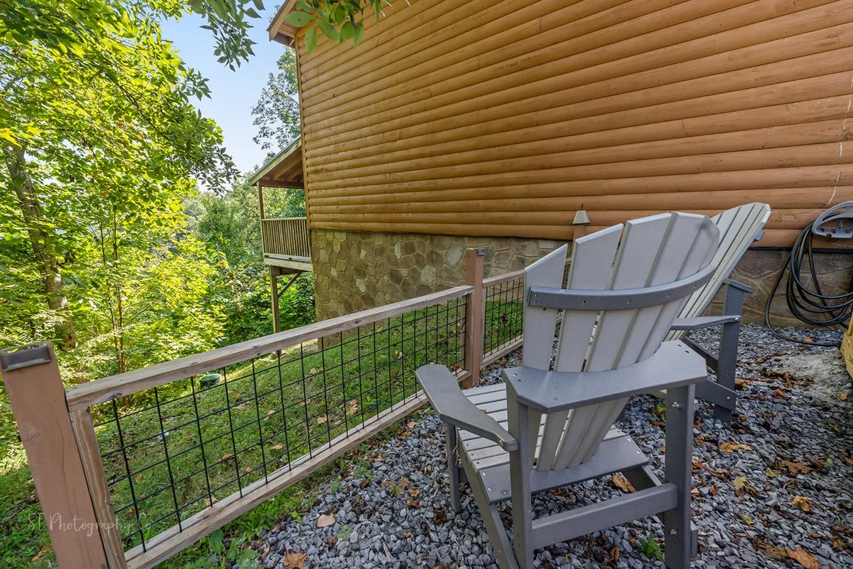 An adirondack chair is positioned on a wooden deck bordered by a rustic stone wall and lush greenery, offering a serene spot for relaxation and socializing. The view hints at tranquil forest surroundings, ideal for quiet moments or late-night chats with friends.